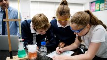 A photo of a small group of middle school students looking over a chemistry chart as their teacher supervises in the background.