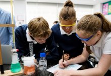 A photo of a small group of middle school students looking over a chemistry chart as their teacher supervises in the background.