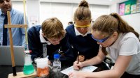 A photo of a small group of middle school students looking over a chemistry chart as their teacher supervises in the background.