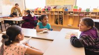A photo of a elementary students smiling and talking to each other as their teacher talks to other students in the background.