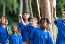A photo of elementary students outside with their teacher, and all the students are pointing and looking up into the trees.