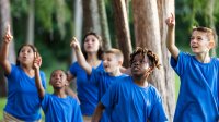 A photo of elementary students outside with their teacher, and all the students are pointing and looking up into the trees.