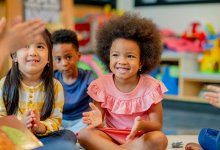 A photo of kindergarteners sitting and clapping with their teacher who has a book folded over their lap.