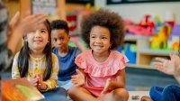 A photo of kindergarteners sitting and clapping with their teacher who has a book folded over their lap.