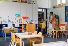 A photograph of a kindergarten teacher greeting excited students as they enter a colorful and decorated classroom.