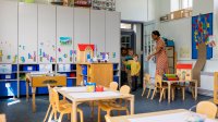 A photograph of a kindergarten teacher greeting excited students as they enter a colorful and decorated classroom.