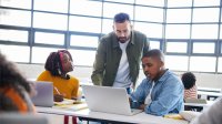Teacher talking to two students as they complete a daily routine on computers