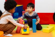 A photo of two preschool students moving to build a structure out of large blocks.