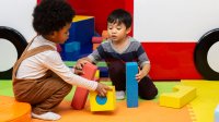 A photo of two preschool students moving to build a structure out of large blocks.