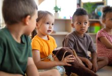 An image of kindergarteners sitting in a circle facing a teacher off-screen. The student in the center is holding a dog plushie.