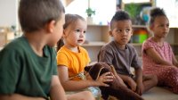 An image of kindergarteners sitting in a circle facing a teacher off-screen. The student in the center is holding a dog plushie.