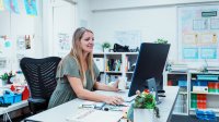 A photo of a teacher at her desk working on a desktop computer.
