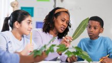 A photo of a teacher and her elementary students holding up leaves as they document them in a journal.