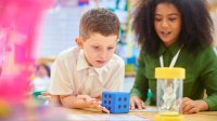 A photo of a teacher assisting an elementary student with a math task involving an hourglass, numbers chart, and die.