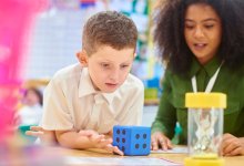 A photo of a teacher assisting an elementary student with a math task involving an hourglass, numbers chart, and die.