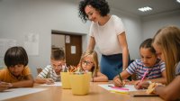 A photo of elementary students engaged in a writing activity as their teacher looks over them and examines her own instructional design.