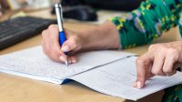 A photo focused on an administrator's hands as they write in a journal at their desk.