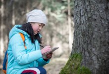 A photo of an elementary student writing on a notepad while observing a tree trunk.