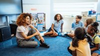 A photo of a teacher sitting in a circle on the floor with their elementary students.