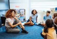 A photo of a teacher sitting in a circle on the floor with their elementary students.