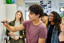 A photo of a high school student starting to write on a white board as his fellow classmates observe.