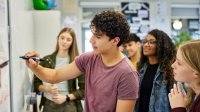 A photo of a high school student starting to write on a white board as his fellow classmates observe.