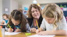 A photo of two elementary students writing as their teacher observes behind them.