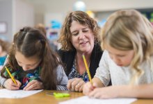A photo of two elementary students writing as their teacher observes behind them.