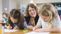 A photo of two elementary students writing as their teacher observes behind them.