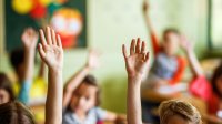 A photo of students in an elementary classroom raising their hands