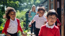 A photo of students excitedly running outdoors as their teacher observes from afar