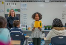 An elementary-aged girl sharing their work in a presentation in front of their class and teacher.