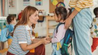 Preschool teacher kneeling down to welcome a student to class
