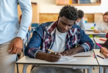 Student working on a paper assignment in class while teacher walks by
